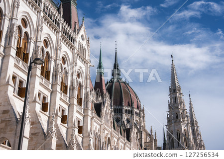 The Hungarian Parliament Building in Budapest features pointed arches, spires, and a central dome, set against a bright blue sky with scattered clouds. The Hungarian Parliament Building in Budapest features pointed arches, spires, and a central dome, set against a bright blue sky with scattered clouds. 127256534