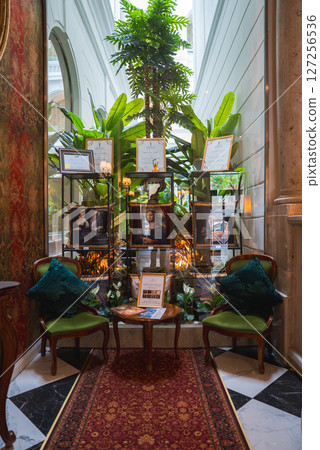 Elegant seating area with vintage chairs, wooden table, and red rug. Glass shelves display awards, framed by tall plants and large windows. 127256536