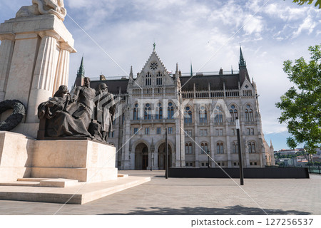 The Hungarian Parliament Building's Gothic Revival architecture stands tall with a detailed memorial statue in the foreground under a partly cloudy sky. 127256537
