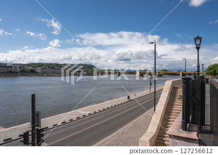 Scenic view of the Danube River with Margaret Bridge in the background, a riverside road, stone staircase, street lamps, and urban greenery. 127256612
