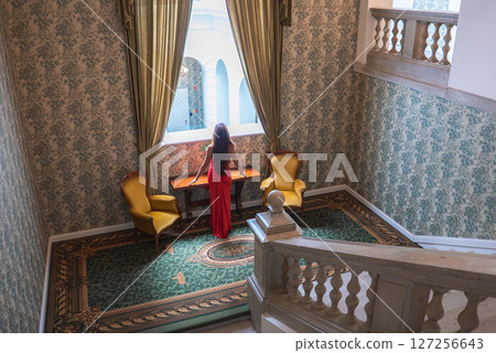 Luxury hotel interior featuring a grand staircase, ornate balustrades, floral wallpaper, and a woman in a red dress by a window with gold curtains. 127256643
