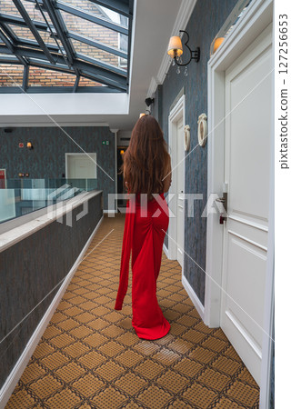A beautiful woman in a red dress walks in a hotel corridor with patterned carpet, white doors, glass railing, skylight, and exposed brick visible above. 127256653