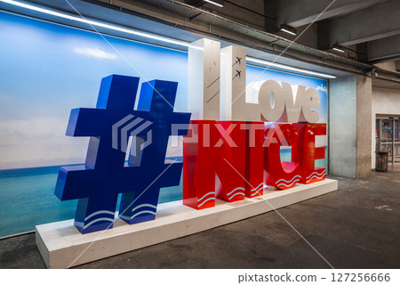 Indoor display of ILoveNice with blue, red, and white design, airplane icons, beach backdrop, and industrial airport setting in Nice, France. 127256666