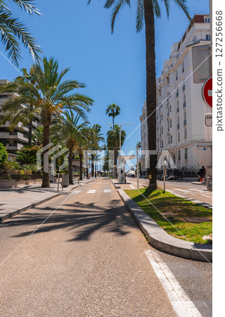 A quiet street in Cannes, France, featuring tall palm trees, elegant buildings, and a white structure resembling the Carlton Hotel under a bright blue sky. A quiet street in Cannes, France, featuring tall palm trees, elegant buildings, and a white structure resembling the Carlton Hotel under a bright blue sky. 127256668