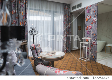 Boutique hotel interior featuring floral curtains, upholstered armchairs, a glass top table, a marble tiled bathroom with a bathtub, and natural light. 127256676