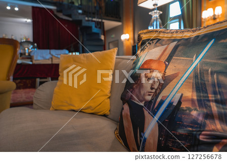 Stylish hotel interior featuring a beige sofa with yellow cushions, a spiral staircase, red curtain, warm lighting, and a dining area in the background. 127256678