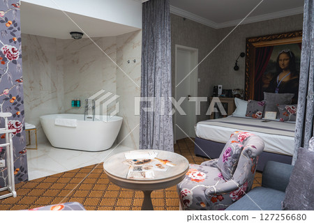 A cozy hotel room featuring a white bathtub in a marble alcove, floral wallpaper, plush furniture, a large bed, and a framed Mona Lisa portrait. 127256680