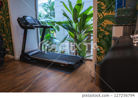 A gym in a luxury boutique hotel featuring a TRX 100 treadmill, tropical wallpaper, wooden flooring, greenery through glass windows, and a water dispenser. A gym in a luxury boutique hotel featuring a TRX 100 treadmill, tropical wallpaper, wooden flooring, greenery through glass windows, and a water dispenser. 127256794