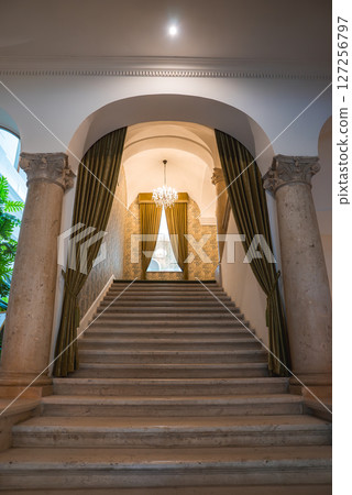 A grand staircase framed by ornate stone columns, green drapes, and a sparkling chandelier in a luxury boutique hotel's refined interior. A grand staircase framed by ornate stone columns, green drapes, and a sparkling chandelier in a luxury boutique hotel's refined interior. 127256797