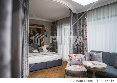 Elegant hotel room featuring a large bed, framed Mona Lisa portrait, floral curtains, upholstered chairs, gray sofa, and natural light from windows and skylight. 127256816