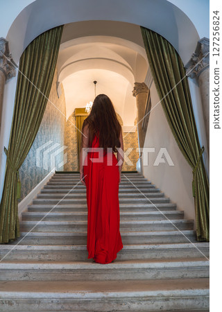 A woman in a red dress ascends a grand staircase with green drapes, ornate columns, and a chandelier, showcasing a luxurious boutique hotel interior. A woman in a red dress ascends a grand staircase with green drapes, ornate columns, and a chandelier, showcasing a luxurious boutique hotel interior. 127256824