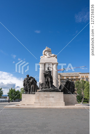 The Tisza Istvan Memorial in Budapest features bronze sculptures and a central statue, set against a blue sky with a crane and historic building visible. 127256836