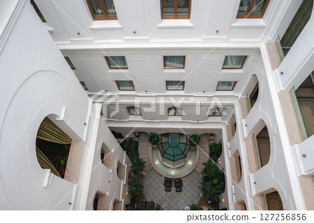 Interior courtyard of a boutique hotel with white facade, arched windows, glass skylight, lush greenery, tiled floor, and balconies from above. 127256856
