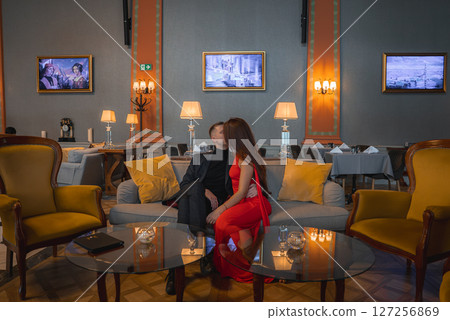 A man in black and a woman in red sit on a gray sofa in a refined hotel interior. Warm lighting, mustard armchairs, and framed artwork enhance the ambiance. A man in black and a woman in red sit on a gray sofa in a refined hotel interior. Warm lighting, mustard armchairs, and framed artwork enhance the ambiance. 127256869
