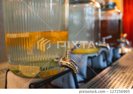 Three glass dispensers with metal spigots hold citrus infused water and mint. Elevated on stands, they rest on white cloths in a warm, upscale setting. 127256905