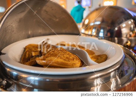 Close up of golden brown pancakes in a stainless steel chafing dish, with warm lighting, reflective surfaces, and a person in a teal hoodie in the background. Close up of golden brown pancakes in a stainless steel chafing dish, with warm lighting, reflective surfaces, and a person in a teal hoodie in the background. 127256906