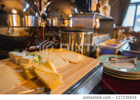 An elegant cheese buffet on a wooden board with fresh herbs, metallic serving domes, and cutlery in a well lit boutique hotel dining area. 127256916