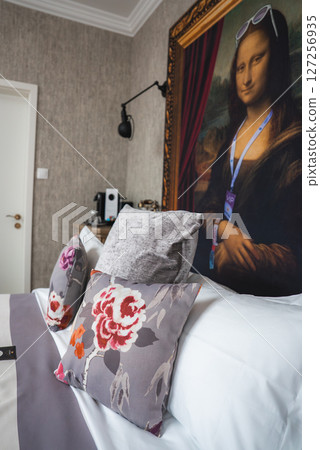 Stylish hotel room featuring a Mona Lisa portrait with sunglasses, floral pillows on a gray bed, a coffee machine, and contemporary decor elements. Stylish hotel room featuring a Mona Lisa portrait with sunglasses, floral pillows on a gray bed, a coffee machine, and contemporary decor elements. 127256935