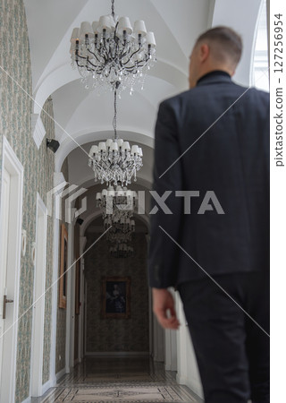 A man in a dark suit walks through a hallway with arched ceilings, ornate chandeliers, patterned wallpaper, and framed artwork in an upscale setting. A man in a dark suit walks through a hallway with arched ceilings, ornate chandeliers, patterned wallpaper, and framed artwork in an upscale setting. 127256954