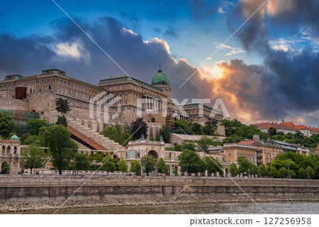 Buda Castle with its green dome and intricate stonework stands on a hill above the Danube River, framed by a tree lined promenade and vibrant clouds. 127256958