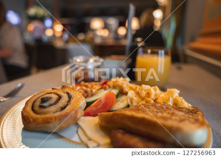 A cinnamon roll, toast, eggs, beans, and vegetables on a plate with orange juice. Warmly lit dining area with elegant decor and blurred figures in the background. A cinnamon roll, toast, eggs, beans, and vegetables on a plate with orange juice. Warmly lit dining area with elegant decor and blurred figures in the background. 127256963
