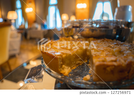 A partially sliced pie with a golden crust and glossy filling on a glass plate, set against a boutique hotel interior with warm lighting and arched windows. A partially sliced pie with a golden crust and glossy filling on a glass plate, set against a boutique hotel interior with warm lighting and arched windows. 127256971