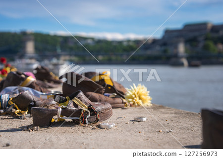 Holocaust memorial iron shoes with ribbons and flowers on the Danube Bank in Budapest, Hungary, with the Chain Bridge and Buda Castle in the background. Holocaust memorial iron shoes with ribbons and flowers on the Danube Bank in Budapest, Hungary, with the Chain Bridge and Buda Castle in the background. 127256973