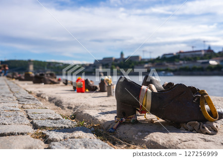 Iron shoes line the Danube riverbank at the Holocaust memorial in Budapest, Hungary, with Buda Castle and city landmarks visible under a partly cloudy sky. Iron shoes line the Danube riverbank at the Holocaust memorial in Budapest, Hungary, with Buda Castle and city landmarks visible under a partly cloudy sky. 127256999