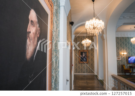 Hallway of a luxury boutique hotel featuring ornate chandeliers, patterned wallpaper, framed portraits, arched doorways, and intricate tile flooring. 127257015