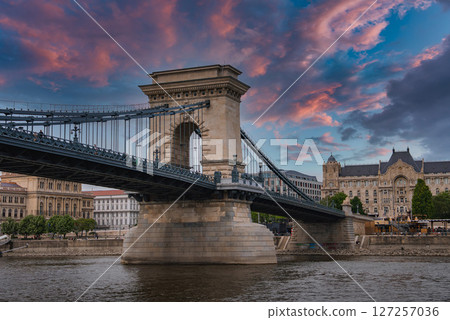 The Szechenyi Chain Bridge spans the Danube River in Budapest under a colorful sunset, with stone towers, suspension cables, and a historic building visible. The Szechenyi Chain Bridge spans the Danube River in Budapest under a colorful sunset, with stone towers, suspension cables, and a historic building visible. 127257036