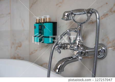A polished chrome vintage style faucet and handheld shower adorn a marble wall with beige and gold veining. Turquoise toiletries add a touch of color. A polished chrome vintage style faucet and handheld shower adorn a marble wall with beige and gold veining. Turquoise toiletries add a touch of color. 127257047