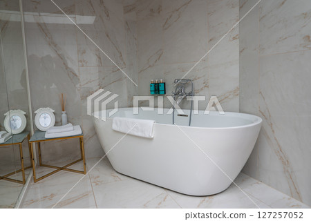 Elegant bathroom featuring a white freestanding bathtub, marble tiles with beige veining, gold framed side table, and decorative mirrors. 127257052