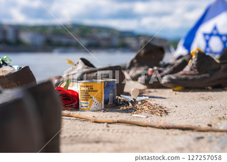 Iron shoes, a red rose at the Holocaust memorial on the Danube Bank memorial in Budapest, Hungary. The Danube River and a blurred Israeli flag are visible in the background. 127257058