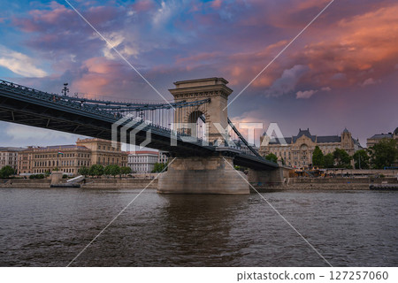 The Szechenyi Chain Bridge spans the Danube River in Budapest, Hungary, with stone towers, suspension cables, and a vibrant sunset sky reflected in the water. 127257060