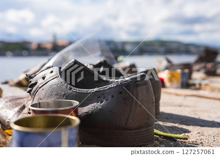 Weathered iron shoes at the Shoes on the Danube Bank Holocaust memorial in Budapest, with candles, flowers, the Danube River, and cityscape in the background. 127257061