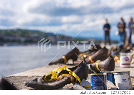 The Shoes on the Danube Bank Holocaust memorial in Budapest features bronze shoes with yellow ribbons, paint cans nearby, and the Danube River in the background. 127257062