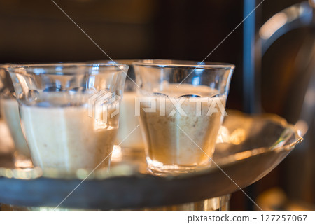 Several clear glass cups with creamy beverages rest on a golden tray under warm lighting, with a blurred elegant interior in the background. Several clear glass cups with creamy beverages rest on a golden tray under warm lighting, with a blurred elegant interior in the background. 127257067