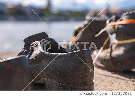 Bronze shoes of the Holocaust memorial line the Danube River in Budapest, with a blurred cityscape and water visible in the background. 127257081
