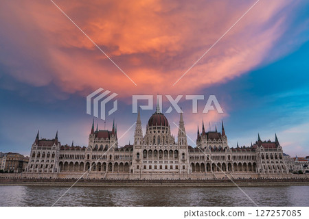 The neo Gothic Hungarian Parliament Building in Budapest, Hungary, stands by the Danube River under a vibrant orange and pink sky with calm reflections. 127257085