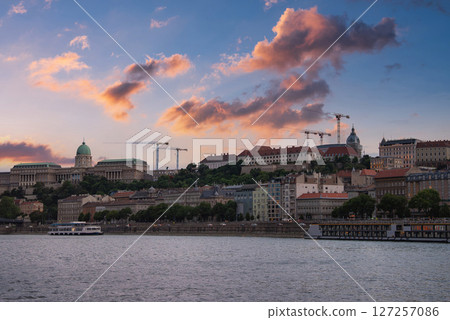 View of Budapest, Hungary, featuring Buda Castle, the Danube River with a boat, Matthias Church, construction cranes, and a sunset sky with clouds. View of Budapest, Hungary, featuring Buda Castle, the Danube River with a boat, Matthias Church, construction cranes, and a sunset sky with clouds. 127257086