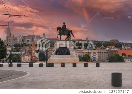 An equestrian statue in Budapest, Hungary, stands on a paved square with the Hungarian Parliament in the background under a sunset sky with a rainbow. 127257147
