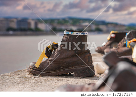 Bronze shoe sculpture with ribbon and Hebrew text at Shoes on the Danube Bank memorial in Budapest, Hungary, with Danube River in the background. Bronze shoe sculpture with ribbon and Hebrew text at Shoes on the Danube Bank memorial in Budapest, Hungary, with Danube River in the background. 127257154