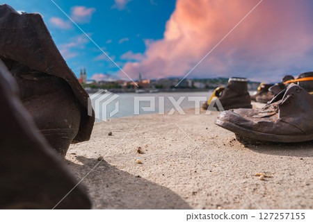 Rusted iron shoes of the Holocaust memorial on the Danube riverbank in Budapest, Hungary, with a cityscape and vibrant pink and orange sky. 127257155