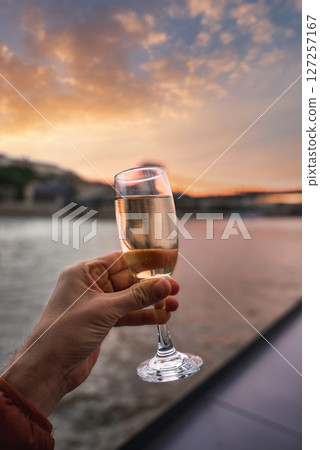 A hand holds a champagne glass by the Danube River at sunset in Budapest, Hungary. Soft orange and pink hues reflect on the water with buildings in view. 127257167