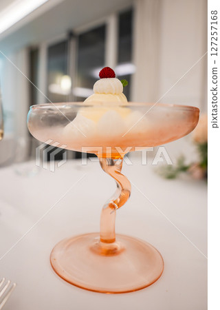 A creamy dessert served in a pink hued twisted stem glass, garnished with a red berry and mint leaf. Set in a modern dining space with large windows. 127257168