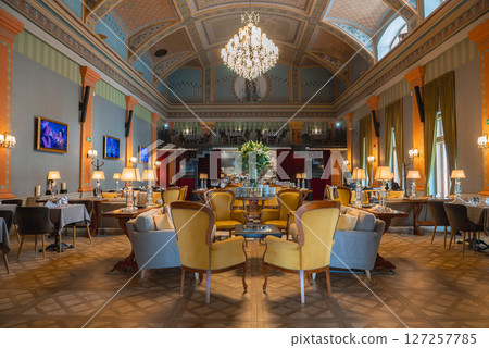 High ceilinged dining area with ornate details, vibrant armchairs, floral centerpieces, framed artwork, tall windows, and a staircase in view. 127257785