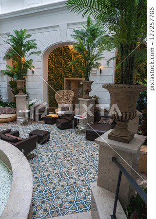 A cozy hotel interior featuring patterned tiled floors, lush greenery, wicker seating, a stone fountain, arched doorway, and tropical wallpaper. 127257786