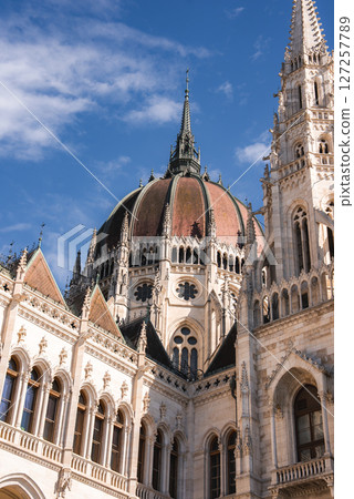 The Hungarian Parliament Building in Budapest features a neo Gothic facade with pointed arches, ornate carvings, and a central dome under a blue sky. 127257789