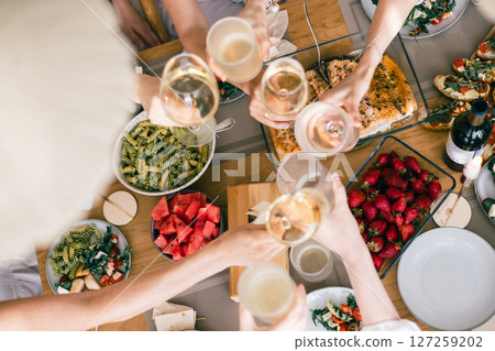 Group of friends toasting with wine over festive table with pasta, salmon, strawberries, watermelon Group of friends toasting with wine over festive table with pasta, salmon, strawberries, watermelon 127259202