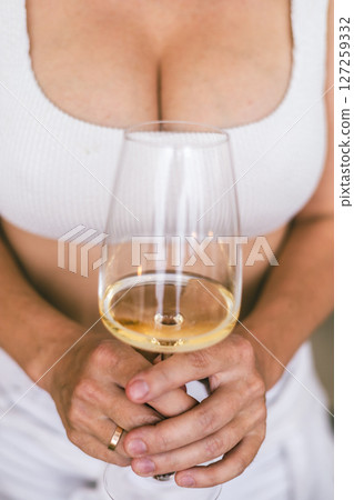 Woman in white top holding a glass of white wine, close-up of hands, chest in soft natural lighting 127259332